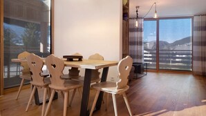 A dining area with a wooden table and chairs, a modern pendant light, and a view through a large window. - Holiday Apartment "Noreia Granda" with Mountain View, Pool, Wi-Fi, Terrace & Balcony (St.Ulrich)