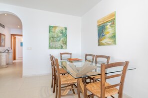 Un coin repas avec une table à plateau de verre entourée de chaises en bois, deux tableaux accrochés aux murs et un couloir menant à une autre pièce.