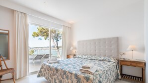 Une chambre avec un grand lit, deux lampes de chevet, une table de nuit en bois, un fauteuil en osier et une vue sur la mer par une porte vitrée.
