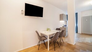 Ein moderner Essbereich mit einem weißen Tisch, vier Stühlen, einem an der Wand befestigten Flachbildfernseher und einer Küche im Hintergrund.