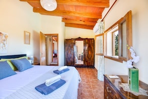 Een slaapkamer met een houten plafond, een bed met blauw-witte beddengoed, een dressoir, een spiegel en een raam met gordijnen.