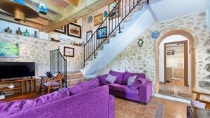A living room with a stone wall, a staircase, a television, and purple sofas. - Pet-friendly Villa Cala Veritat with Mountain View, Pool, A/C, Wi-Fi, Garden, Terrace & Balcony (inca)
