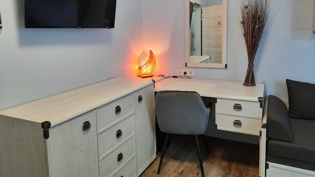 Een moderne kamer met een flatscreen televisie aan de muur, een bureau met lades, een stoel en een nachtkastje met een vaas droogbloemen.