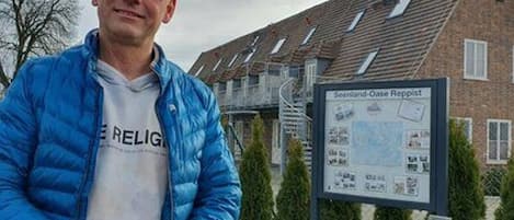 Um homem com casaco azul em frente a um edifício com uma placa que diz "Seenland-Oase Roppist".
