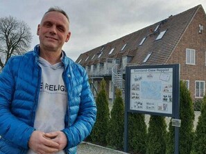 Un homme vêtu d’une veste bleue se tient devant un bâtiment sur lequel on peut lire l’inscription « Seenland-Oase Roppist ».