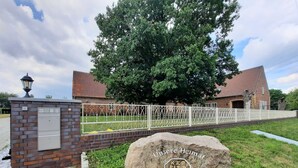 A building with a brick facade, a white metal fence, and a large tree in front. - Zimmer 5 - Seenland-oase Reppist (Senftenberg)