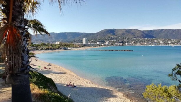 Una playa costera con agua azul cristalina, una orilla arenosa y colinas y edificios al fondo.