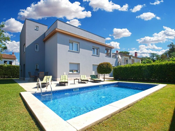 Ein modernes, zweistöckiges Haus mit Swimmingpool, Terrasse und Liegestühlen sowie einem gepflegten Rasen.