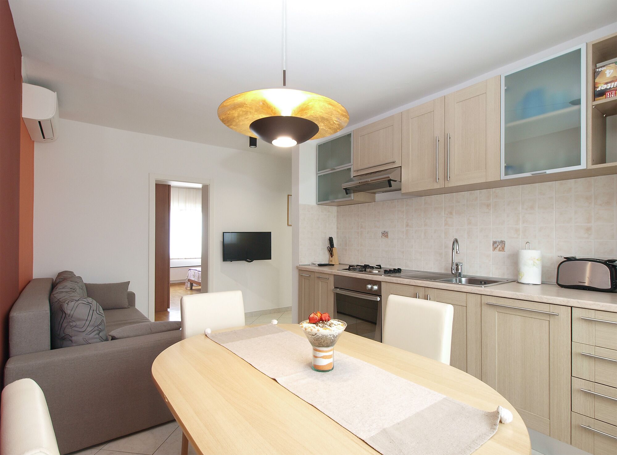 A modern kitchen with a dining area, a TV mounted on the wall, and a sofa in the living room.