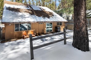Un chalet dont le toit est recouvert de neige, avec une terrasse en bois et un grand arbre au premier plan.