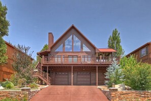 A two-story log cabin with a balcony, a garage, and a stone retaining wall. - Luxury Log Home with Mountain Views! Quiet Family Neighborhood! Gameroom! Firepit! (Big Bear City)