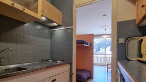 A compact kitchen with a sink, stove, and wooden cabinets. A bed is visible in the adjacent room, and a window offers a view of trees. - Cozy studio with snow front view, close to chairlift and shops (Le Dévoluy)