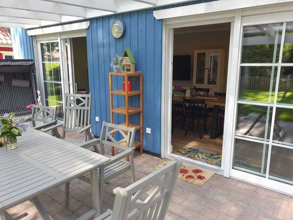 An outdoor patio with a table and chairs, a wooden shelf with decorative items, and a blue wall with a window.