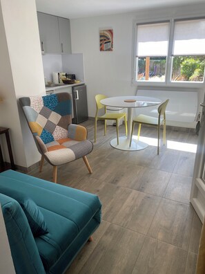 Een moderne woonkamer met een blauwgroene bank, een fauteuil met een patroon, en een kleine eettafel met gele stoelen. Achtergrond zichtbaar: een keuken met een koelkast en kasten.