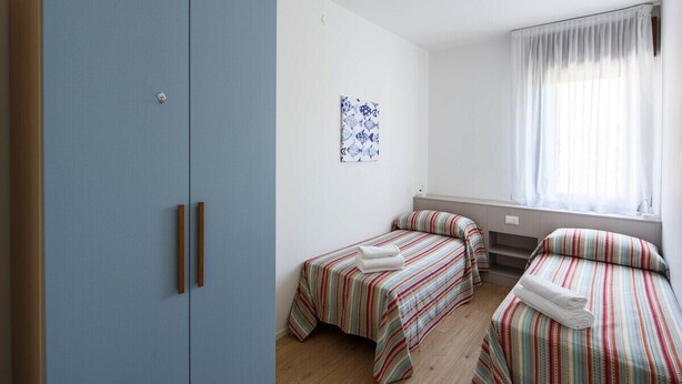 A bedroom with two single beds, a wardrobe, a window with curtains, and a wall-mounted artwork.