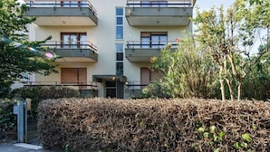 A multi-story residential building with balconies, a central entrance, and a hedge in the foreground.