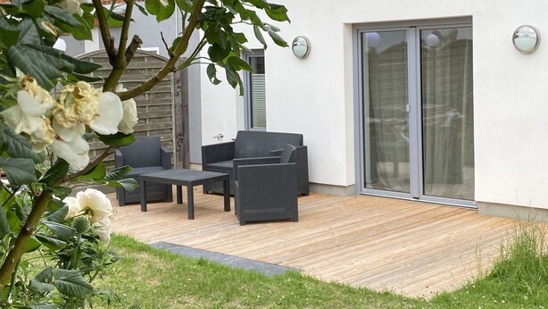 A patio with outdoor furniture, a wooden deck, and a white building with a brick wall and large sliding glass doors. - Idyllisches Ferienhaus in Ostseenähe (Dorf Mecklenburg OT Rambow)