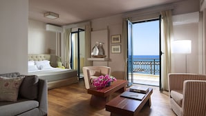 Habitación de hotel con cama, sofá, una mesita de madera, una silla y un balcón con vista al mar.
