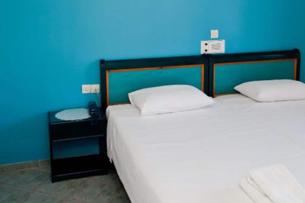 A neatly made bed with white linens and pillows, a bedside table with a lamp, and a power outlet on the wall.