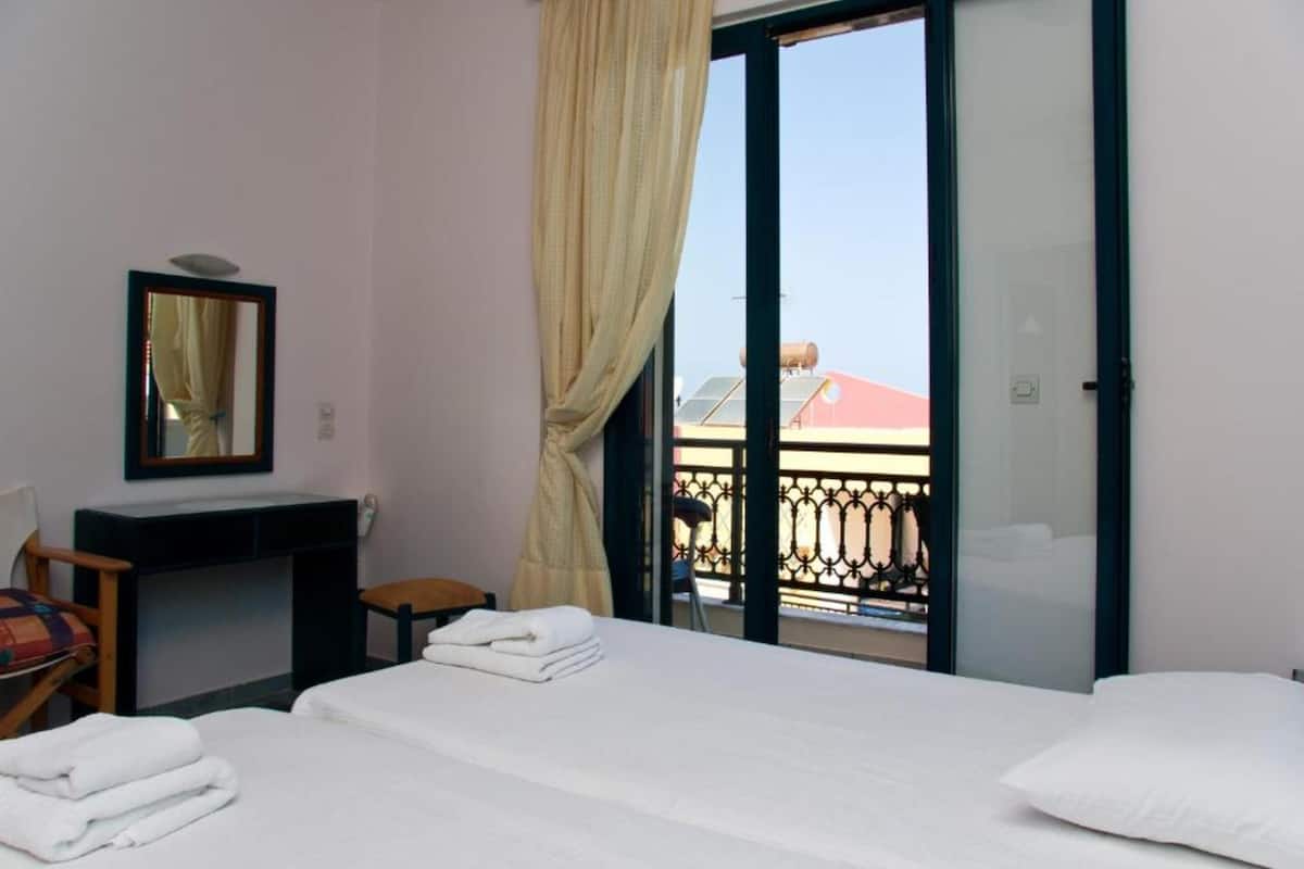 A hotel room with two beds, a desk, a chair, a mirror, and a balcony with a view of buildings.
