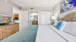 A hotel room with a bed, a TV on a wooden dresser, a bathroom with a mirror, and a painting on the wall. - Surf Beach 304 - Spectacular Sunsets - Beach Front (Treasure Island)