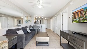 A modern living room with a sofa, coffee table, TV, and a dining area with chairs and a table. - Surf Beach 304 - Spectacular Sunsets - Beach Front (Treasure Island)