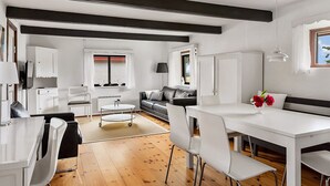 Et moderne køkken med hvide skabe, et spiseområde med et hvidt bord og stole, en sort sofa og et vindue med udsigt.
