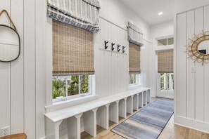 Un pasillo con bancos blancos, ventanas amplias con persianas y un espejo decorativo.
