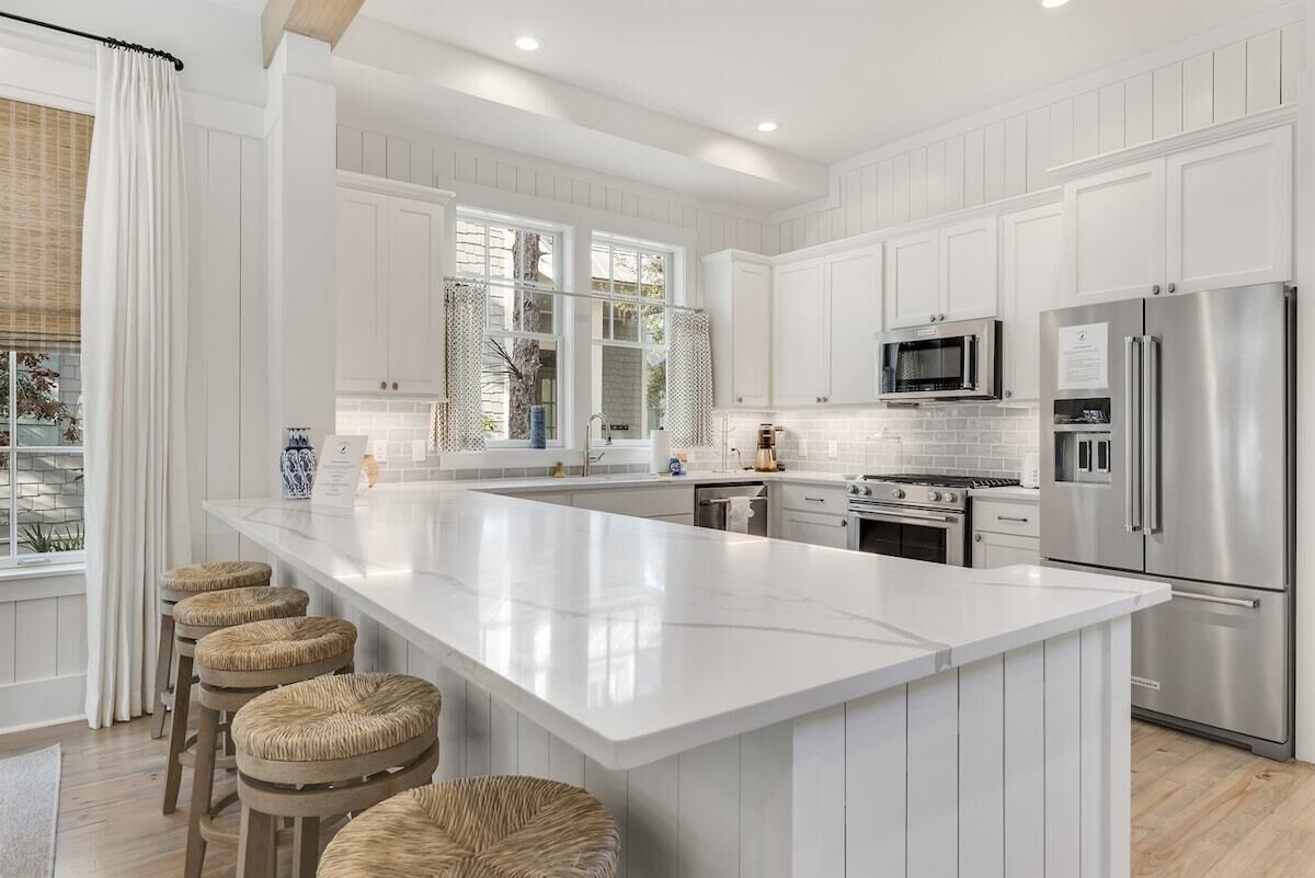 A modern kitchen with white cabinetry, a large island, bar stools, stainless steel appliances, and a view of the outdoors.