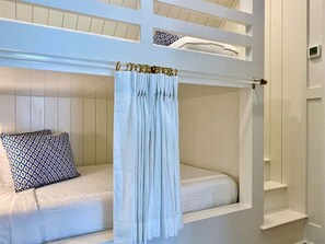Uma cama de solteiro com dossel, um travesseiro estampado e uma parede branca com prateleira embutida.
