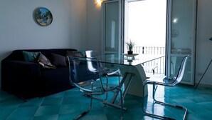 Un espace de vie moderne avec une table et des chaises en verre, un canapé foncé et une vue sur un balcon extérieur, accessible par une porte vitrée.