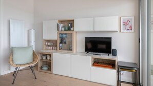 Uma sala de estar moderna com uma televisão de tela plana, uma poltrona de vime e um aparador de vidro com livros e objetos decorativos.