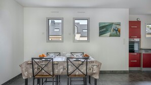 Un coin repas avec une table dressée pour quatre personnes, deux fenêtres, une cuisine rouge et un tableau accroché au mur.