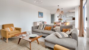 Una sala de estar moderna con un sofá, un sillón y una mesa de centro. Se observa una cocina al fondo con armarios blancos y una zona de comedor.
