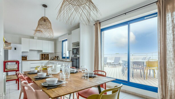 Un comedor moderno con una mesa de madera dispuesta para seis personas, rodeada de sillas coloridas. Un amplio ventanal ofrece vistas de la terraza y el cielo.