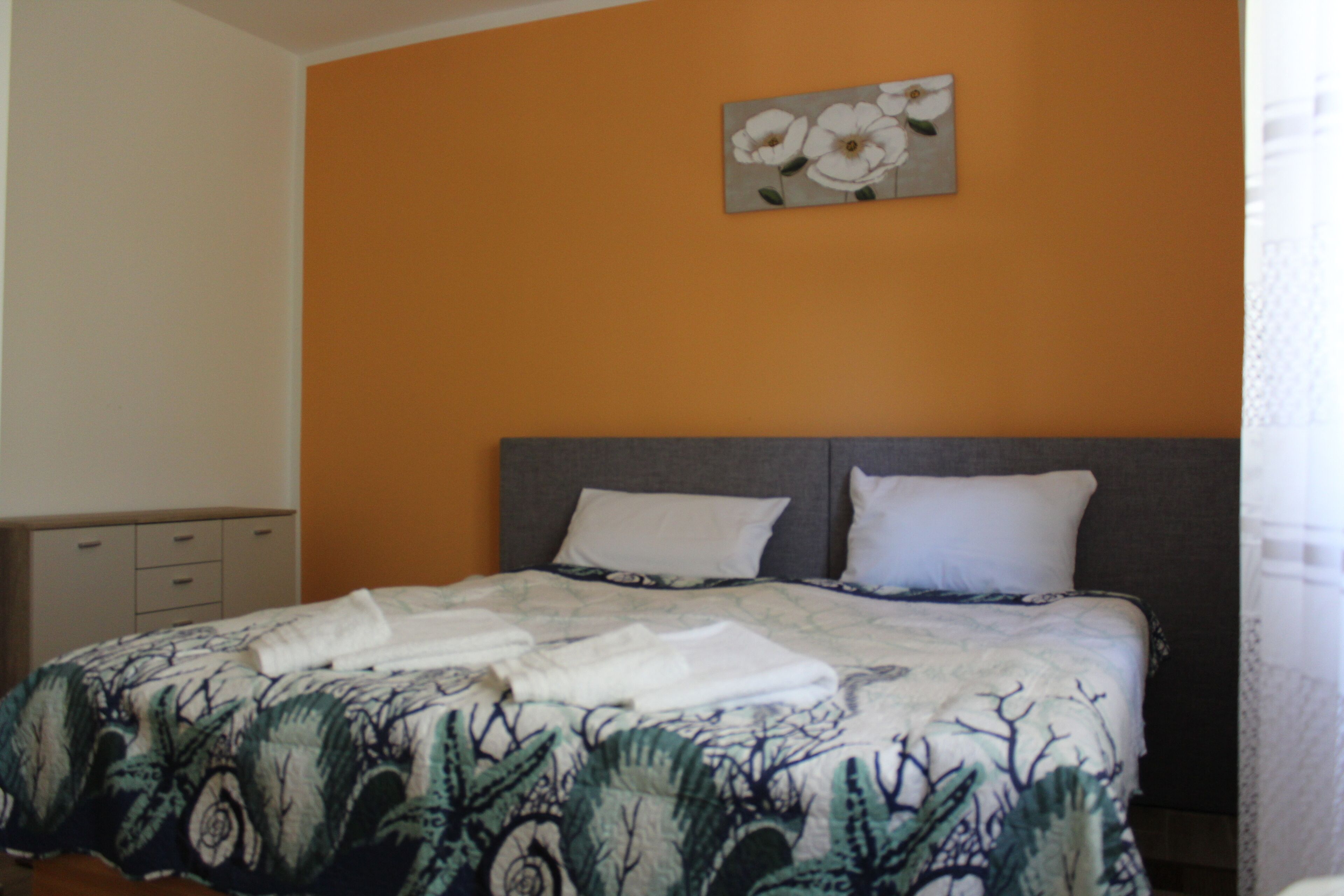 A bedroom with an orange wall, a bed with a patterned bedspread, two pillows, and a painting of flowers.