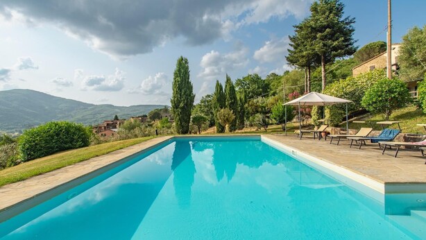 A rectangular swimming pool with a clear blue water surface, surrounded by a paved deck. There are lounge chairs and a covered seating area with a table. The pool is set against a backdrop of trees and a mountainous landscape.