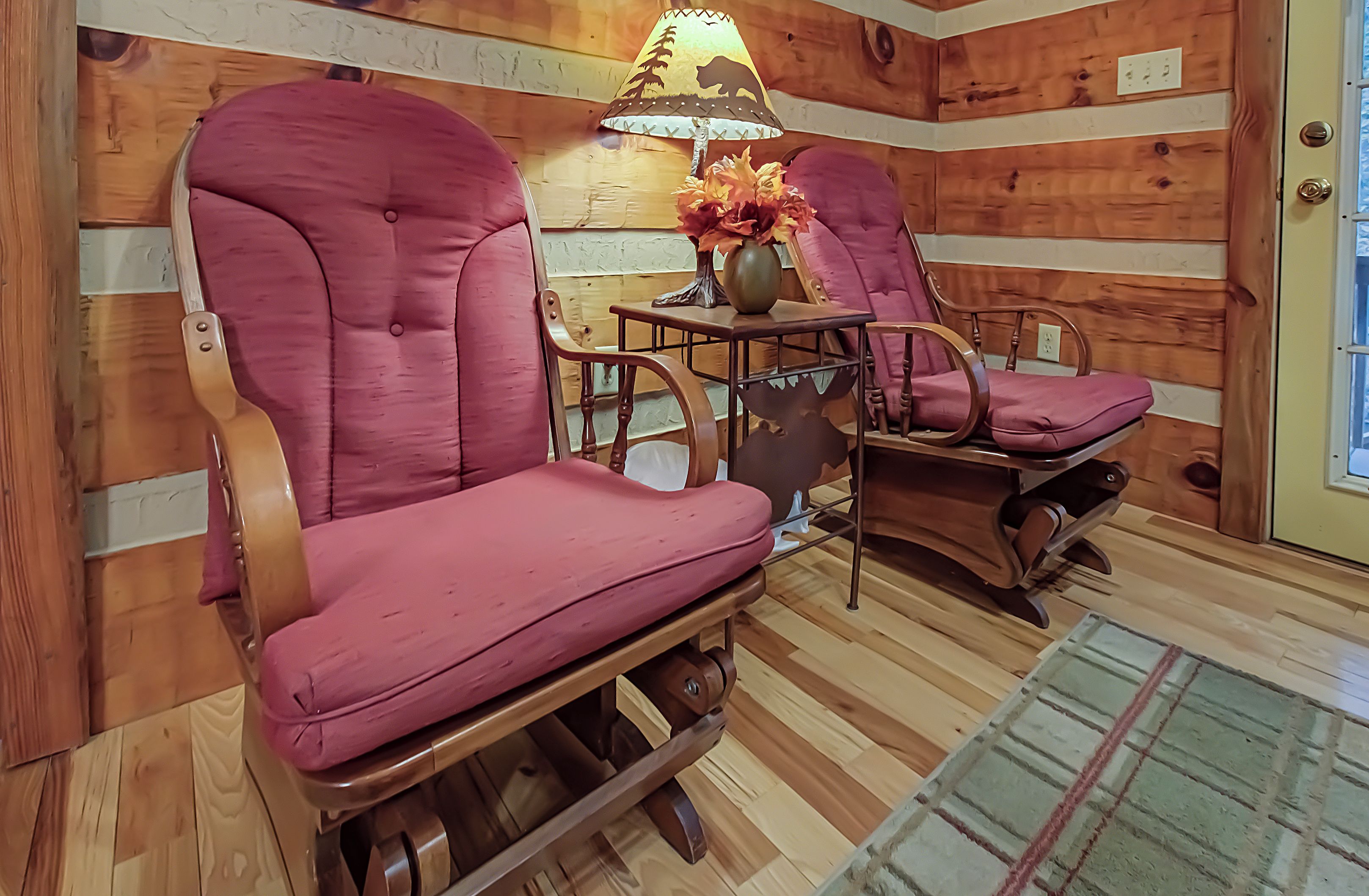 A cozy room with wooden walls and a wooden floor. There are two rocking chairs with purple cushions and a table with a lamp and flowers.