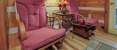 A cozy room with wooden walls and a wooden floor. There are two rocking chairs with purple cushions and a table with a lamp and flowers.