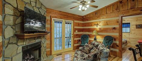 Ein gemütliches Cabin-Interieur mit Kamin aus Stein, Deckenventilator und großem Fenster mit Blick auf Bäume.