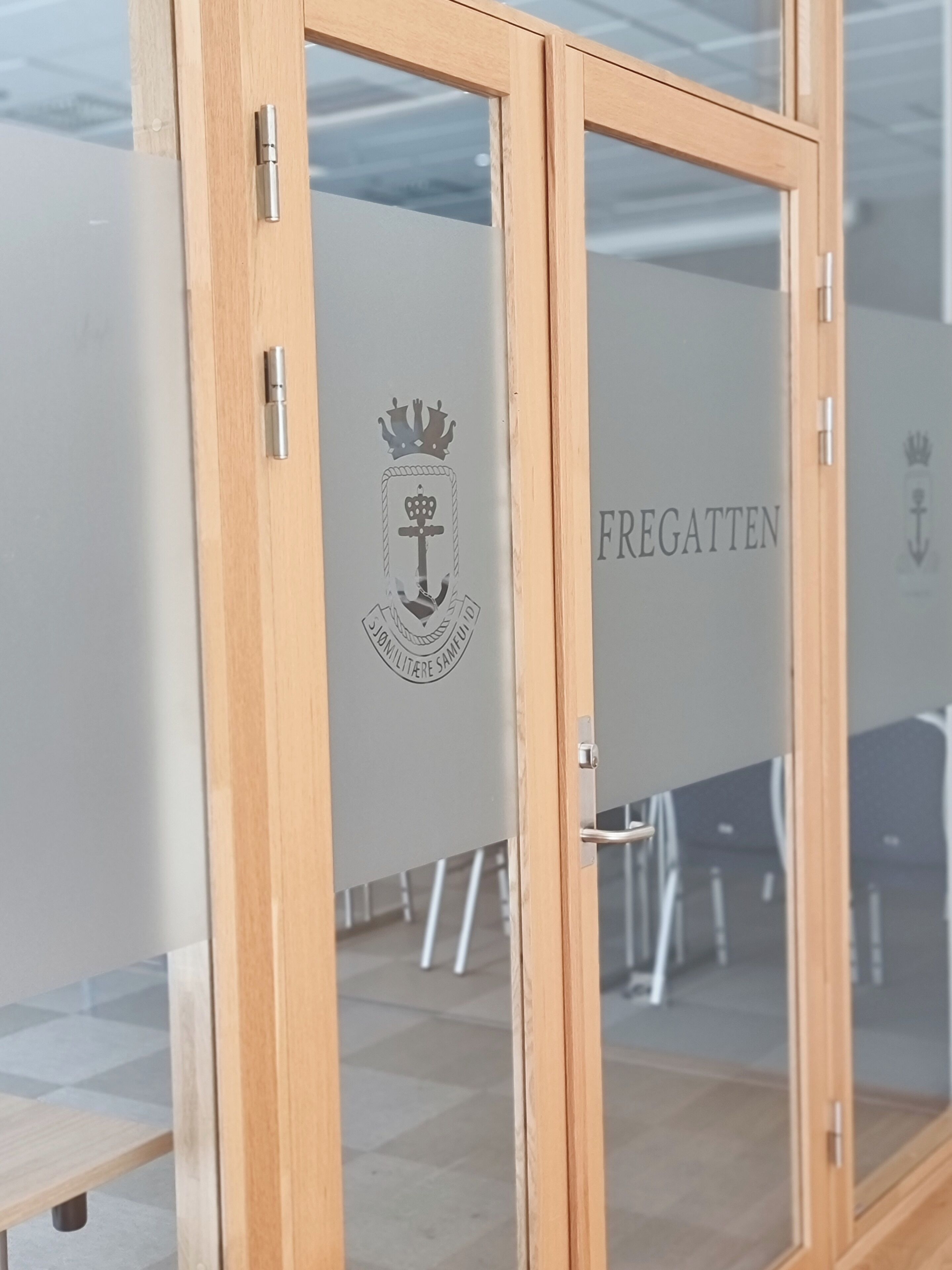 A room with a wooden door and a glass wall. The wall has a logo with a crown and anchor, and the word "FREGATTEN" is visible.