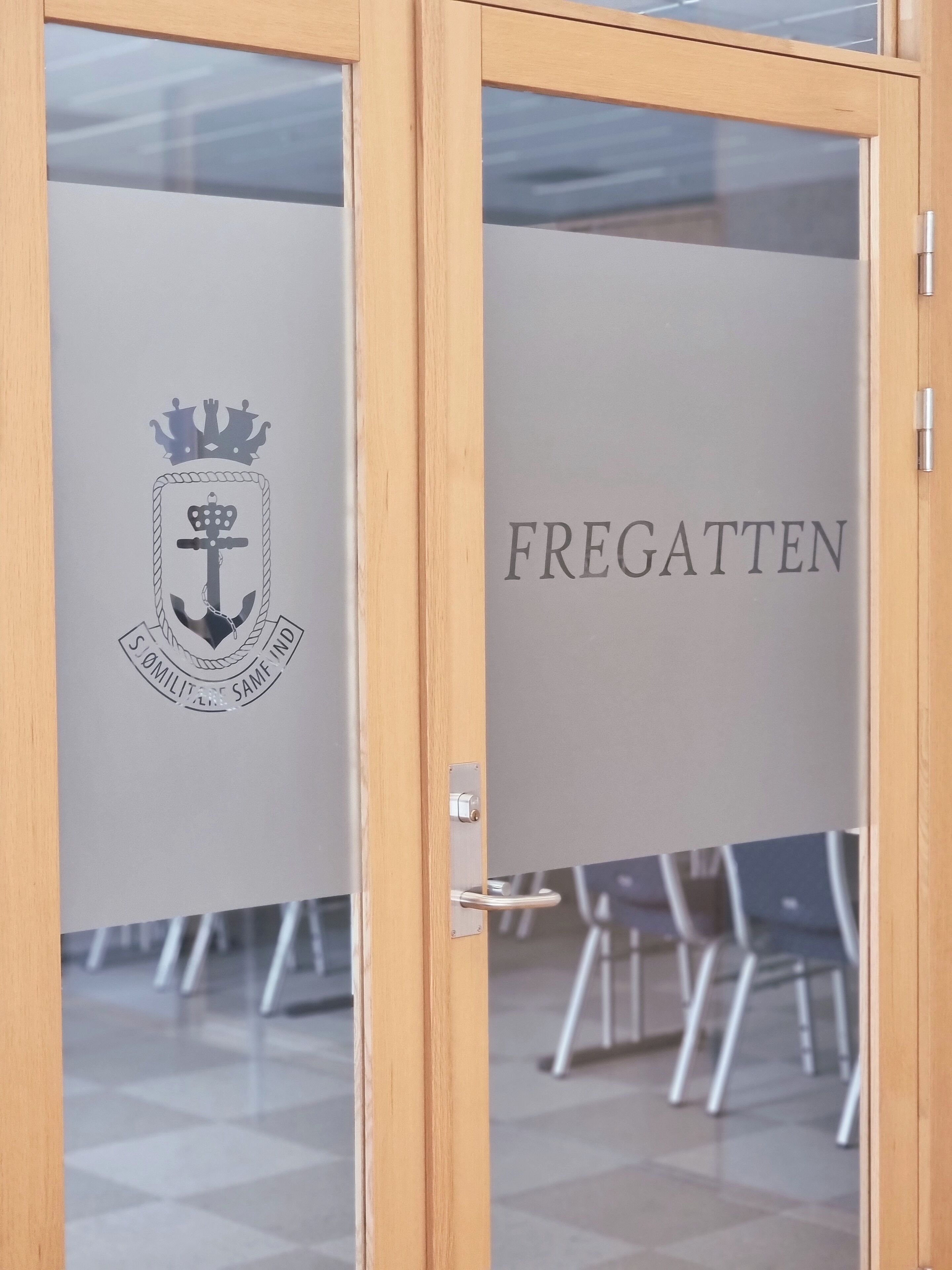 A wooden-framed door with frosted glass panels. The left panel displays a crest with a crown, anchor, and the text "SOMILITERS SAMFIND". The right panel reads "FREGATTEN".