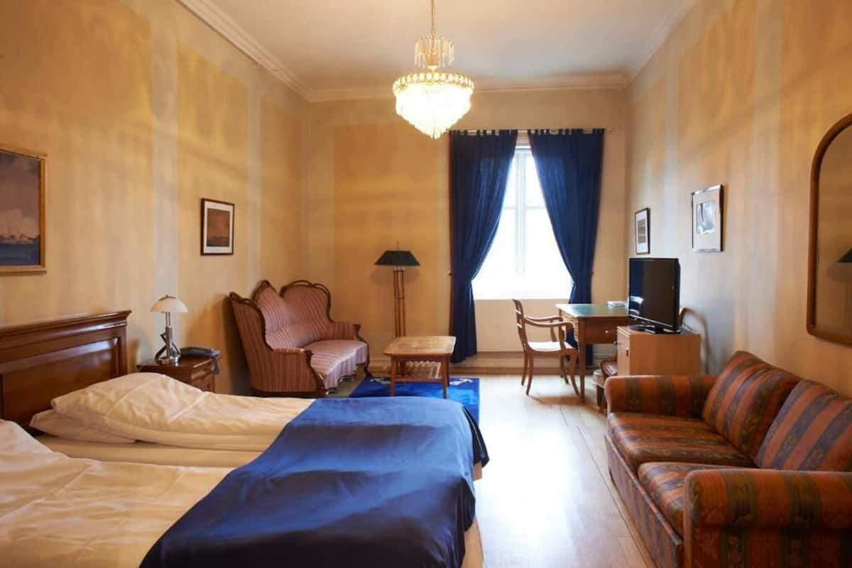 A hotel room with two beds, a sofa, a desk, a chair, a television, and a chandelier.