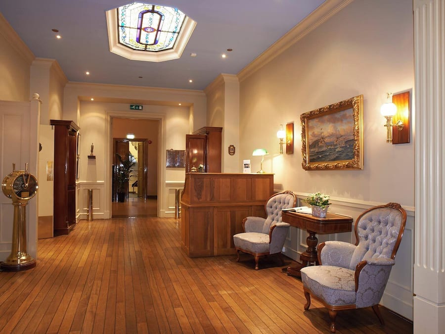 Un vestíbulo de hotel con suelo de madera, una recepción, dos sillones tapizados, un cuadro en la pared y un amplio ventanal con diseño de vitral.