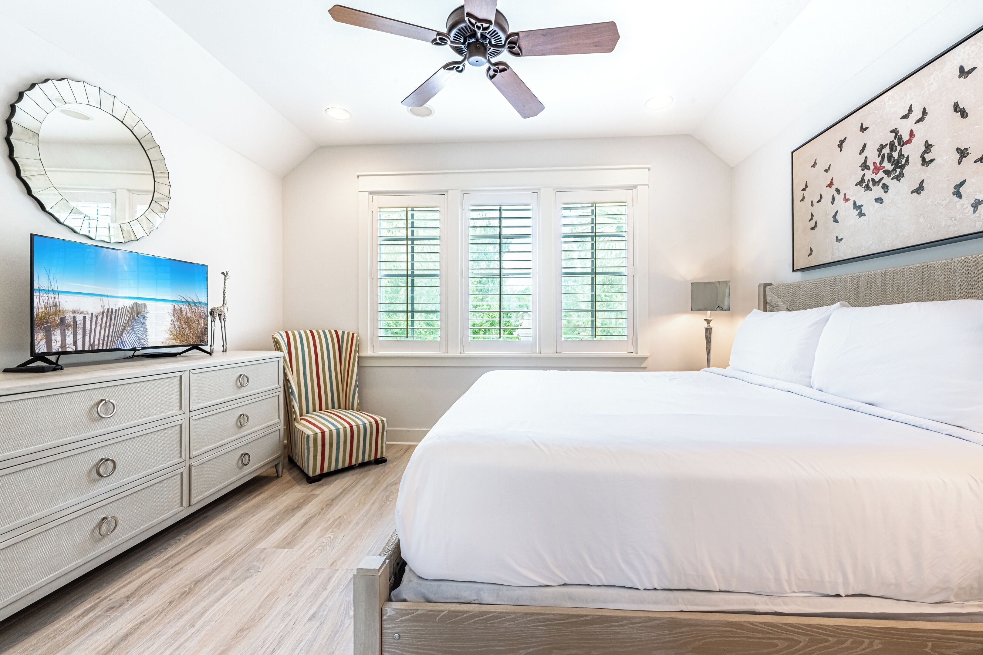 Une chambre à coucher comprenant un lit, une commode, une chaise, un ventilateur de plafond, une fenêtre avec des stores et un tableau accroché au mur.