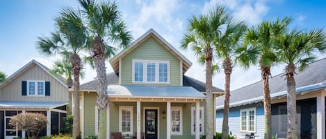 A two-story house with a green exterior, a front porch with chairs, and palm trees.