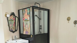 A bathroom with a black shower enclosure, a white sink with a mirror, and a small vanity. There is a blue chair with a floral cushion and a blue rug on the floor.