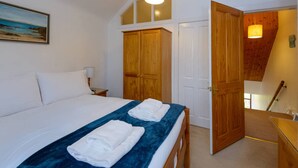 A bedroom with a bed, a nightstand, a wooden wardrobe, and a door leading to another room. - Dunlin - 1 Bedroom Seaside Apartment (North Berwick)