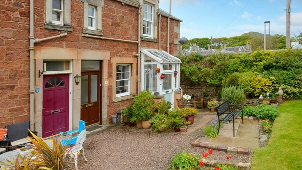 A two-story stone house with a red door, a conservatory, and a garden with potted plants and a bench. - Dunlin - 1 Bedroom Seaside Apartment (North Berwick)