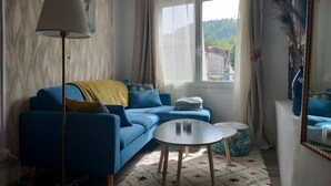 Una sala de estar con un sofá azul, una mesita redonda pequeña, una lámpara y una ventana con cortinas.